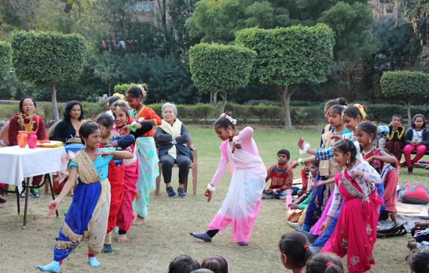 A group of children and adults in a garden surround a girl who is dancing in the middle.